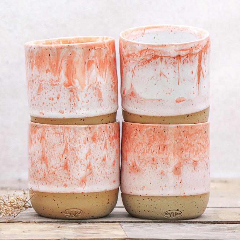 May include: Four ceramic mugs stacked, featuring a speckled, marbled design in coral and white. The mugs have a natural, speckled tan base and a cylindrical shape. The overall aesthetic is modern and handcrafted.