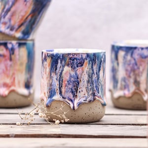 Könnte beinhalten: Ein Keramikbecher mit einer marmorierten Glasur in Blau-, Rosa- und Orangetönen, die über eine gesprenkelte, hellbeige Basis fließt. Der Becher hat eine zylindrische Form mit einer glatten, glänzenden Oberfläche oben und einer matten Textur unten.