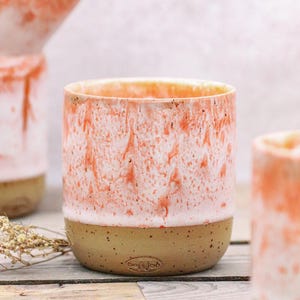 May include: A ceramic cup with a speckled tan base and a coral and white marbled design. The cup has a cylindrical shape and a smooth, unglazed base. The interior is a light orange color.
