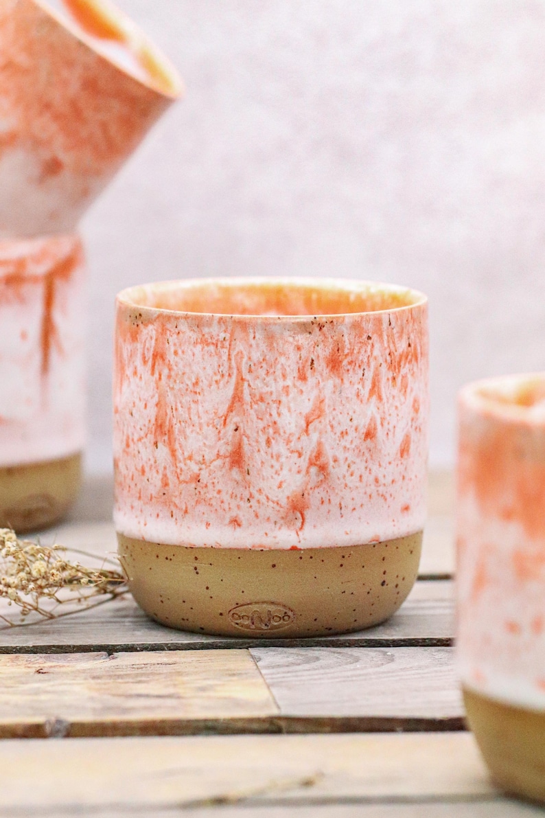 May include: A ceramic cup with a speckled tan base and a coral and white marbled design. The cup has a cylindrical shape and a smooth, unglazed base. The interior is a light orange color.