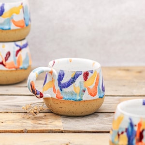 Peut inclure: Mug en céramique blanche avec une base tachetée beige et une anse arrondie. Le mug est décoré de coups de pinceau abstraits colorés dans les tons de bleu, orange, jaune et rouge. Plusieurs mugs sont empilés sur une surface en bois.