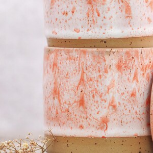 May include: Two ceramic mugs stacked, featuring a speckled tan base and a white and coral marbled design. The mugs have a rounded shape and a smooth finish. The mugs are part of a pottery collection.
