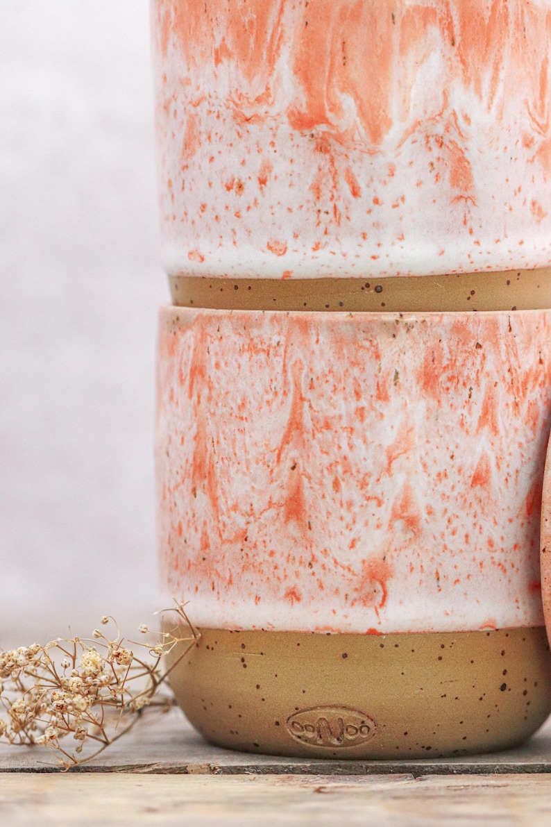 May include: Two ceramic mugs stacked, featuring a speckled tan base and a white and coral marbled design. The mugs have a rounded shape and a smooth finish. The mugs are part of a pottery collection.