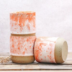 May include: Three ceramic tumblers with a speckled, marbled design in white and coral. The base of each tumbler is a speckled tan color. One tumbler is lying on its side, while the other two are stacked.