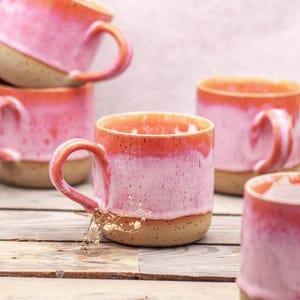 Könnte beinhalten: Eine Sammlung von Keramikbechern mit einer gesprenkelten beigen Basis und einer leuchtenden rosa-orangen Ombré-Glasur. Jeder Becher hat einen stabilen Henkel und einen glatten, abgerundeten Rand. Die Becher sind gruppiert angeordnet.
