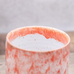 May include: A ceramic mug with a speckled, coral-colored exterior and a white interior. The rim of the mug has a darker coral hue. The mug is part of a set, with a second mug visible in the background. The mugs appear handmade.