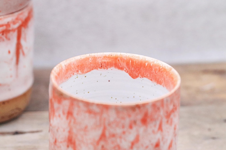 May include: A ceramic mug with a speckled, coral-colored exterior and a white interior. The rim of the mug has a darker coral hue. The mug is part of a set, with a second mug visible in the background. The mugs appear handmade.