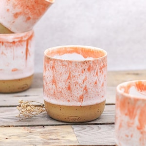 May include: A collection of ceramic mugs with a unique design. The mugs feature a speckled, tan base and a white and coral-colored glaze that creates a marbled effect. The mugs are cylindrical and appear to be handmade.