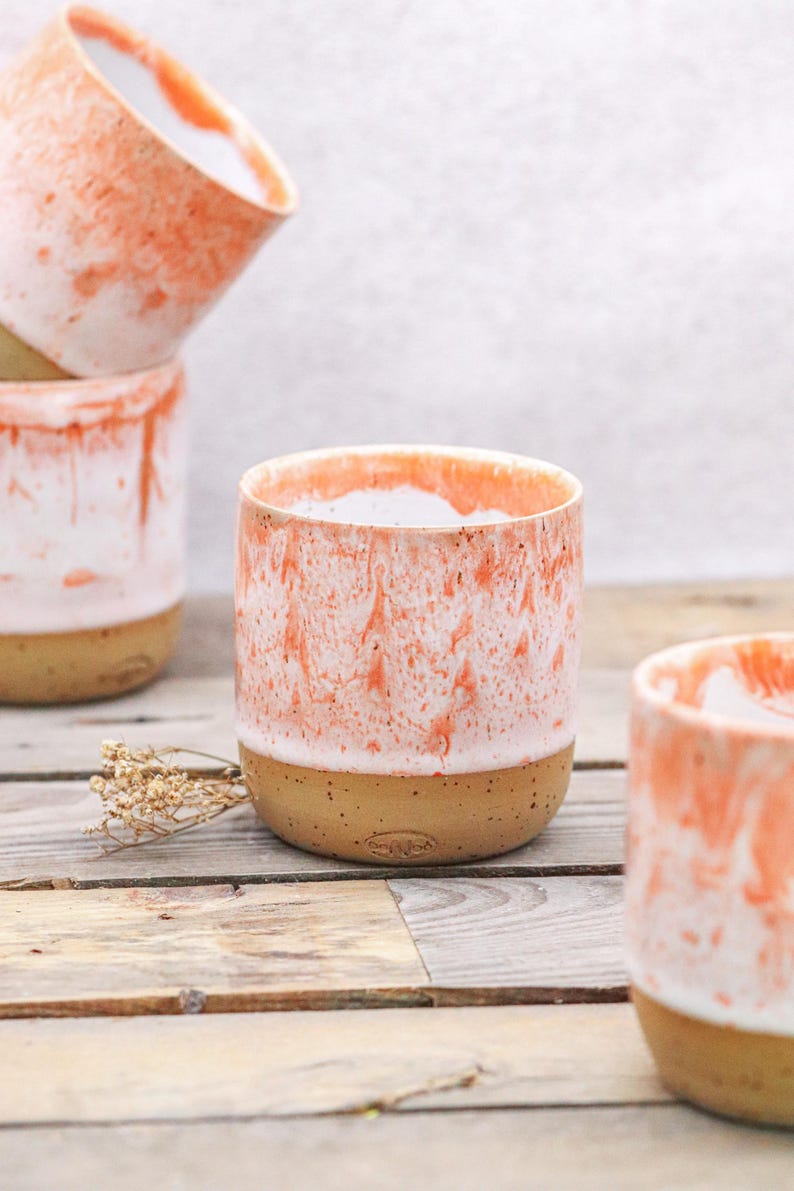 May include: A collection of ceramic mugs with a unique design. The mugs feature a speckled, tan base and a white and coral-colored glaze that creates a marbled effect. The mugs are cylindrical and appear to be handmade.