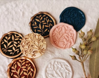 May include: A collection of wax seals in various colors, including black, navy, pink, and gold. Each seal features a detailed botanical design of leaves and branches. The seals are arranged on a white, textured surface.