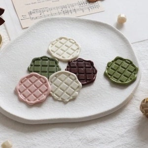 Puede incluir: Seis sellos de cera de diferentes colores con un patr&oacute;n de cuadr&iacute;cula en un plato blanco. Los sellos son en tonos de marr&oacute;n, verde, rosa y blanco.