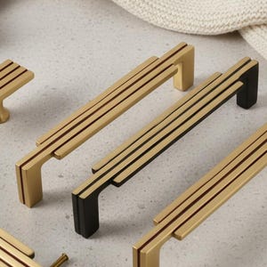 May include: A collection of gold and black cabinet hardware, including handles and a knob. The handles have a linear design with three parallel lines. The hardware is displayed on a light-colored surface.