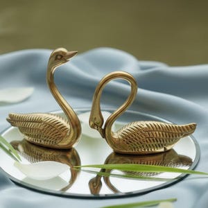 May include: Two gold swan ornaments, with detailed feather patterns, are presented on a small, circular mirror. The swans are set against a backdrop of light blue fabric, with scattered white petals and green blades of grass.