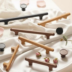 May include: An assortment of wooden and black cabinet hardware, including handles and knobs, displayed on a white satin surface. The handles vary in color from light wood to dark brown. Decorative elements include rose petals and small flowers.