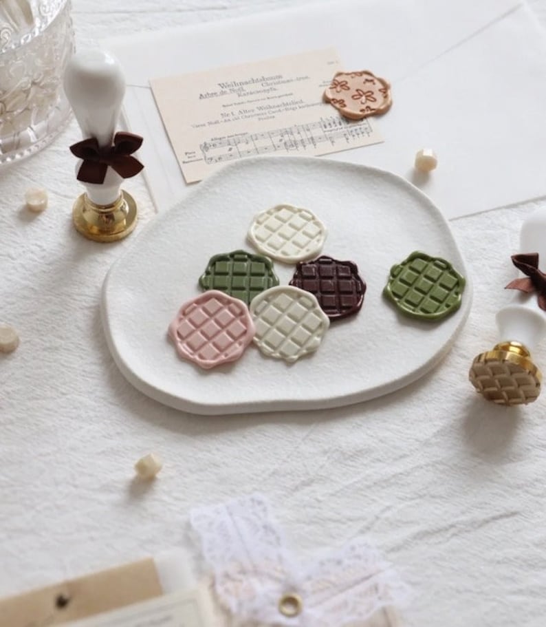 Puede incluir: Seis sellos de cera de diferentes colores con un patr&oacute;n de cuadr&iacute;cula en un plato blanco. Los sellos son en tonos de marr&oacute;n, verde, rosa y blanco.