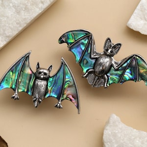 Puede incluir: Dos broches de murciélago plateados con alas iridiscentes en tonos verdes, azules y morados. Los murciélagos tienen detalles finos. Los broches están sobre una superficie beige con detalles de piedra blanca, sugiriendo un tema gótico o de Halloween.