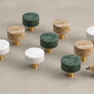 May include: A collection of nine circular cabinet knobs featuring flat, polished stone tops in white, beige, and dark green marble, each mounted on a small, cylindrical brass-toned metal base. The knobs are arranged in a loose grid formation.