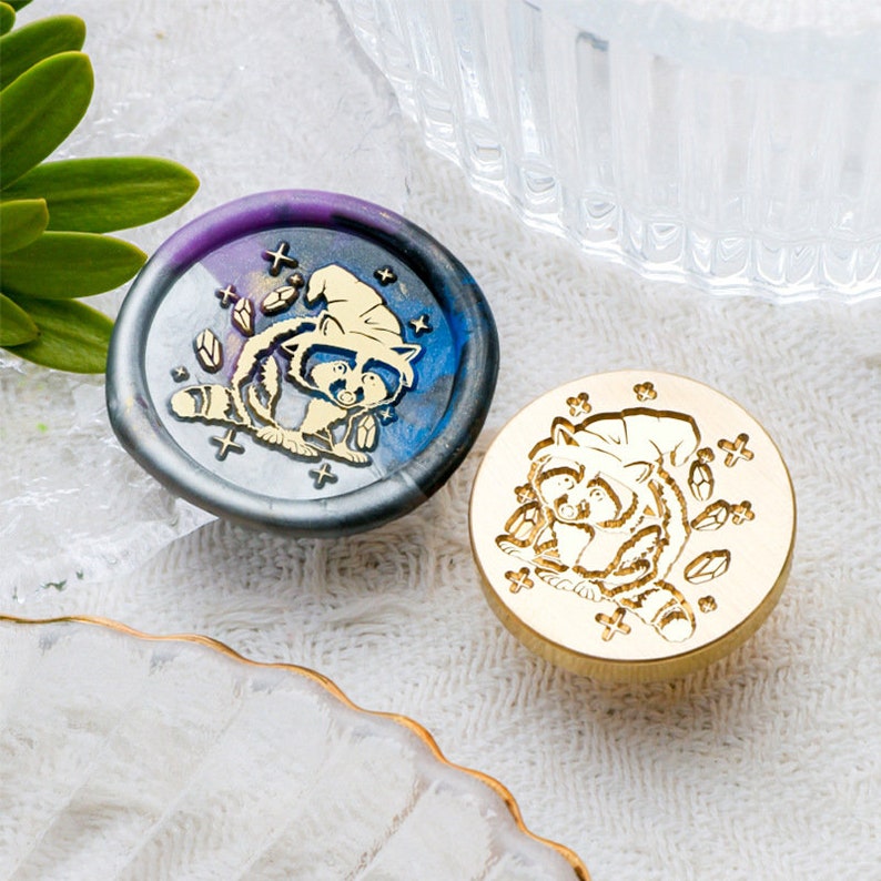 May include: Two wax seals featuring a raccoon wearing a witch's hat. The seal on the left is a dark blue and purple color with gold accents. The seal on the right is a solid gold color.