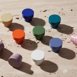 Op de afbeelding: Een verzameling kleurrijke kastknoppen in verschillende tinten, waaronder geel, blauw, blauwgroen, oranje, groen, paars en wit. De knoppen zijn gerangschikt op een lichtgekleurd, getextureerd oppervlak, met schaduwen.