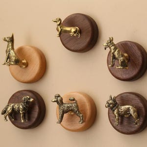 以下が含まれることがあります： 犬をテーマにした装飾的な壁掛けフックのコレクション。 各フックは、木製のディスクに取り付けられた詳細な真鍮製の犬の置物を特徴としています。 犬はさまざまな品種で、ディスクは明るい木材または暗い木材です。