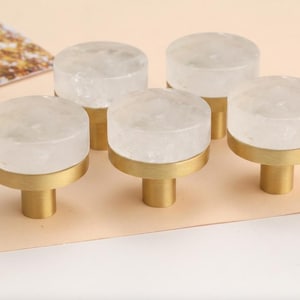 Pode incluir: Puxadores de armário com um topo circular transparente e uma base dourada. Os puxadores são apresentados numa superfície de cor pêssego claro. Os puxadores são provavelmente para móveis.