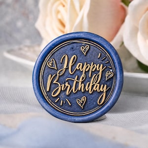 May include: A navy blue wax seal with gold lettering that reads "Happy Birthday." The seal is round with a raised border and features small heart designs. The background includes a white plate and soft pink and white flowers.