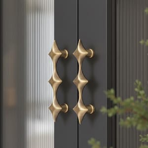 以下が含まれることがあります： スターバーストデザインのアンティーク真鍮製ドアハンドル2つ。ハンドルは濃いグレーのドアに取り付けられており、背景にはテクスチャードガラスパネルと緑があります。ハンドルはユニークな幾何学的な形状をしています。