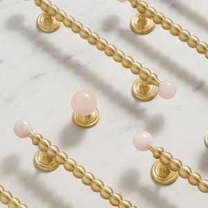 Puede incluir: Primer plano de varios tiradores y pomos dorados para armarios con detalles de piedras preciosas rosas. Los tiradores tienen un diseño de cuentas y los pomos son redondos. El herraje está dispuesto sobre una superficie de mármol blanco.