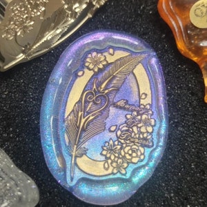 Pode incluir: Um pingente oval de vidro azul e dourado com um design gravado detalhado de uma pena, um cora&ccedil;&atilde;o e flores. O texto "Memory of a Princess" est&aacute; gravado no vidro.