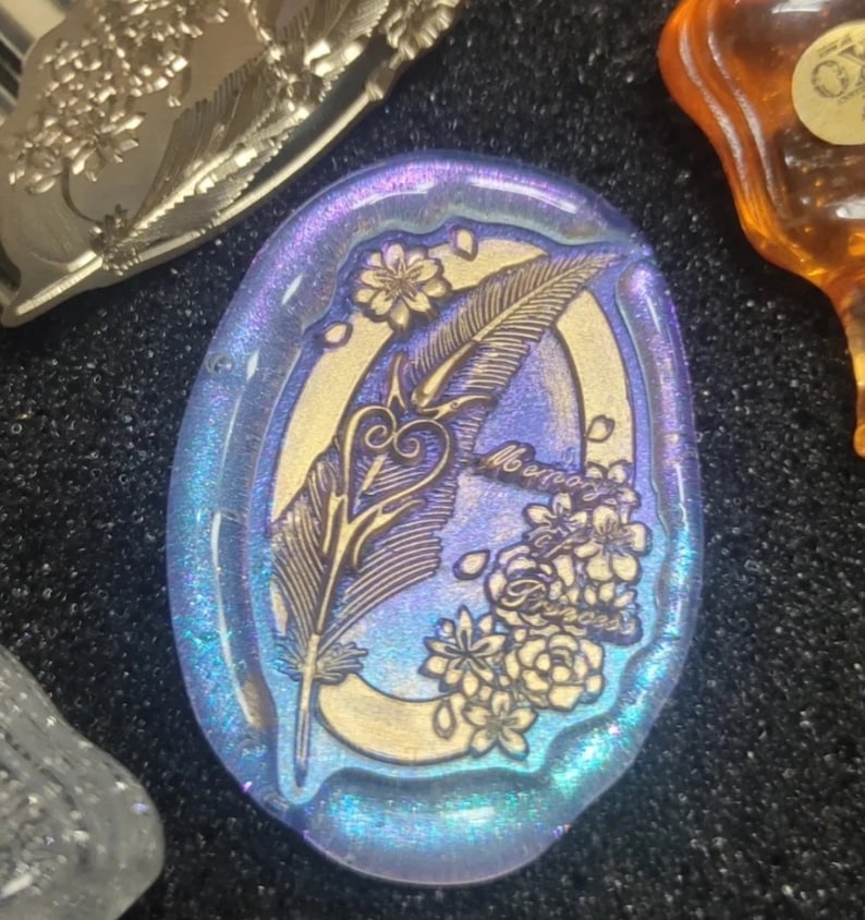 Pode incluir: Um pingente oval de vidro azul e dourado com um design gravado detalhado de uma pena, um cora&ccedil;&atilde;o e flores. O texto "Memory of a Princess" est&aacute; gravado no vidro.