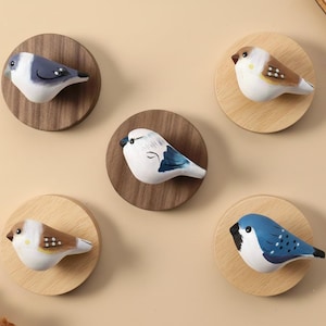 May include: Five decorative wooden bird ornaments, each fixed to a circular wooden base. The birds are painted in shades of blue, white, and brown. The bases are made of light or dark wood. A charming home decor item.