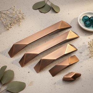 以下が含まれることがあります： さまざまなサイズの幾何学的なブロンズキャビネットプル。プルは、2色の仕上げが施されたモダンで角張ったデザインです。ハードウェアは明るい色の表面に配置されています。