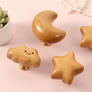May include: Wooden cabinet knobs shaped like a crescent moon, two stars, and a smiling cloud. The knobs are light brown with brass bases. A small succulent plant in a white pot and a pearl bracelet are also in the image.