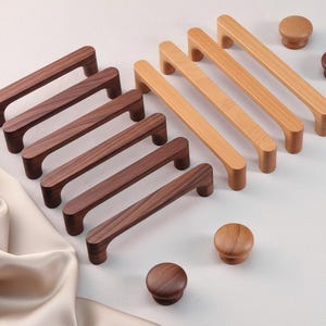 May include: A selection of wooden cabinet handles and knobs in various shades of brown. The handles are rectangular with a curved grip, and the knobs are circular. The hardware is displayed on a white surface, with a satin fabric in the background.