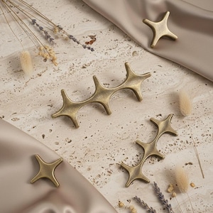 May include: A collection of star-shaped, antique brass cabinet pulls and knobs. The hardware is arranged on a textured, light-colored surface with a satin fabric and dried flowers.
