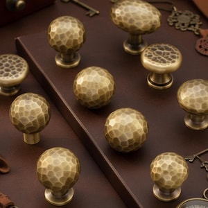 May include: A collection of antique brass cabinet knobs and pulls with a hammered texture. The knobs are various shapes, including round and mushroom-shaped, and are displayed on a dark brown surface. Decorative keys and a compass are also visible.