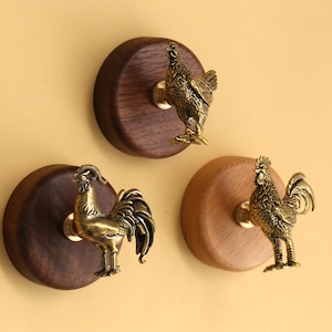 May include: Three decorative wall hooks featuring brass rooster figurines mounted on wooden discs. The hooks are in varying shades of brown and gold, with a warm, inviting aesthetic. The roosters are detailed and add a touch of rustic charm.