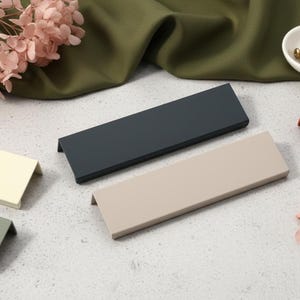 May include: A selection of contemporary cabinet pulls in shades of grey, beige, and terracotta. The rectangular pulls have a small lip. A small white bowl with brass screws and dried pink flowers are also visible.