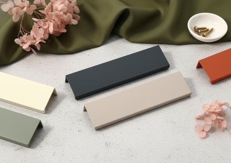 May include: A selection of contemporary cabinet pulls in shades of grey, beige, and terracotta. The rectangular pulls have a small lip. A small white bowl with brass screws and dried pink flowers are also visible.