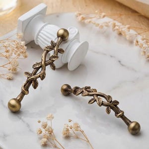 May include: Two antique brass cabinet pulls with a leaf and vine design. Each pull has a spherical end and a decorative center with intertwined leaves. The pulls are displayed on a marble-like surface with a white decorative column in the background.
