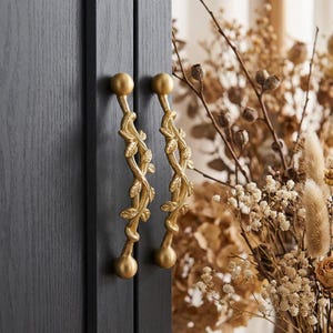 Könnte beinhalten: Zwei goldfarbene Schrankgriffe mit einem Blatt- und Rankenmuster. Die Griffe sind an einer dunkelgrauen Schranktür befestigt. Die Griffe haben oben und unten eine Kugel. Der Hintergrund umfasst getrocknete Blumen.