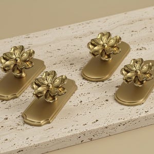 Puede incluir: Cuatro tiradores dorados para armarios con diseño floral. Cada tirador presenta un pomo en forma de flor montado sobre una placa trasera rectangular. Los tiradores están dispuestos sobre una superficie clara y texturizada.