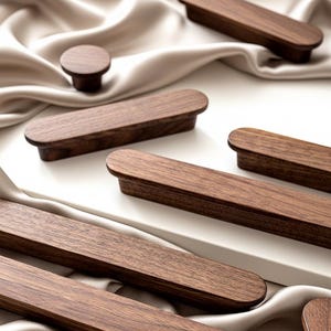 Op de afbeelding: Een verzameling houten meubelbeslag, waaronder verschillende rechthoekige handgrepen en een ronde knop. Het beslag heeft een rijke bruine kleur en lijkt van massief hout te zijn. De items zijn gerangschikt op een wit oppervlak en een crèmekleurige stof.