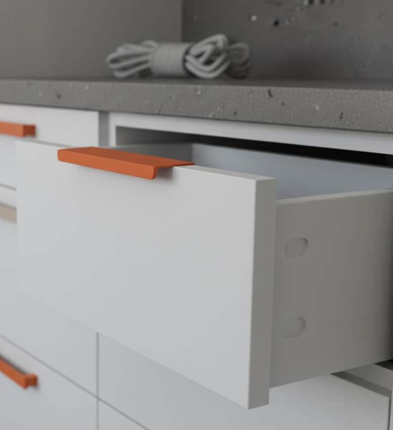 May include: White cabinet with an open drawer and an orange handle. The drawer is part of a modern kitchen or bathroom cabinet set. A rolled-up grey rope sits on the countertop above the cabinet.