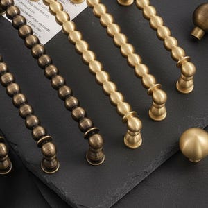 Puede incluir: Primer plano de varios tiradores y pomos de latón para armarios. Los tiradores tienen un diseño de cuentas y están en varias etapas de oxidación. Los pomos son esféricos. El hardware se muestra sobre una superficie oscura.