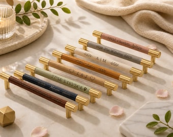 Puede incluir: Una colección de siete tiradores de cajón con agarres cilíndricos de aspecto de cuero en varios colores, incluyendo marrón, azul marino, salvia, tostado, crema y gris, con extremos de metal dorado y grabados láser personalizados.
