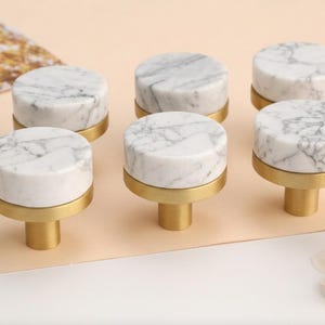 May include: Six marble cabinet knobs with gold-toned bases and stems. The knobs have a circular shape with a white and gray veined pattern. The gold bases add a touch of elegance to the hardware.