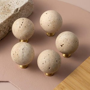 以下が含まれることがあります： ベージュ色の石で作られた、テクスチャ加工された表面を持つ5つの球形のキャビネットノブ。各ノブは金色のベースに取り付けられています。長方形の石と木の表面も画像にあります。