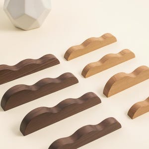 May include: A collection of wooden pen rests in various shades of brown and tan. Each rest has a curved, wave-like design, perfect for holding pens or pencils. A white geometric vase is in the background.