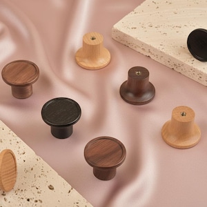 May include: A collection of wooden cabinet knobs in various shades of brown and natural wood tones. The knobs have a round top and a cylindrical base. Some are dark brown, some are light brown, and some are black.
